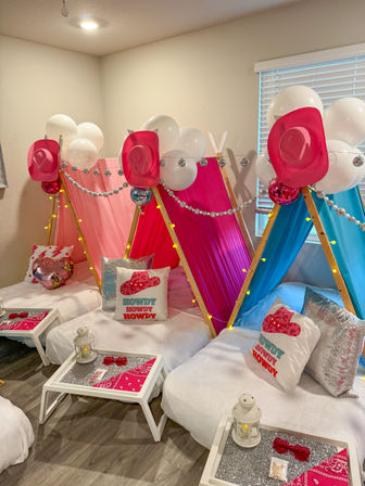 Indoor kids' sleepover setup with three pink and blue mini teepee tents, white balloons, disco balls, pink cowboy hats, 'Howdy' pillows, fairy lights and bedside trays with lanterns.