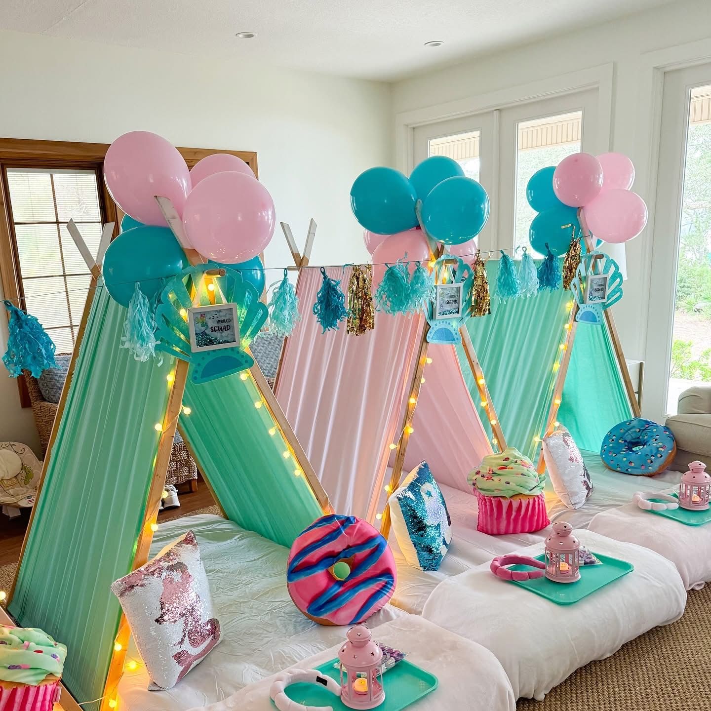Indoor kids' slumber party setup in a bright living room with pastel mini teepee tents in mint and pink, string lights, teal and pink balloons, tassel garlands, donut and cupcake pillows, and individual snack trays with lanterns.