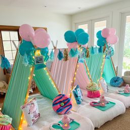 Indoor kids' slumber party setup in a bright living room with pastel mini teepee tents in mint and pink, string lights, teal and pink balloons, tassel garlands, donut and cupcake pillows, and individual snack trays with lanterns.
