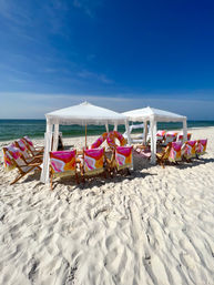 Sunny white-sand beach with two white fringe cabanas, wooden lounge chairs draped in colorful retro pink‑orange towels, inflatable ring, turquoise sea and clear blue sky.