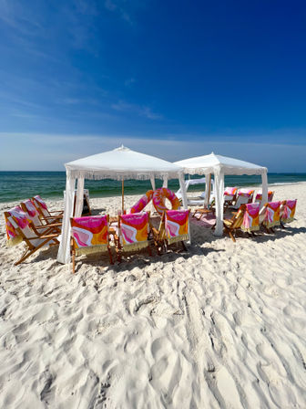 Sunny white-sand beach with two white fringe cabanas, wooden lounge chairs draped in colorful retro pink‑orange towels, inflatable ring, turquoise sea and clear blue sky.