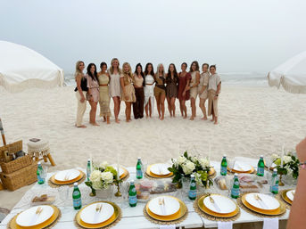Group of women in neutral outfits posing on a white-sand beach behind a boho-chic picnic setup with gold plates, floral centerpieces, fringe umbrellas — seaside celebration.
