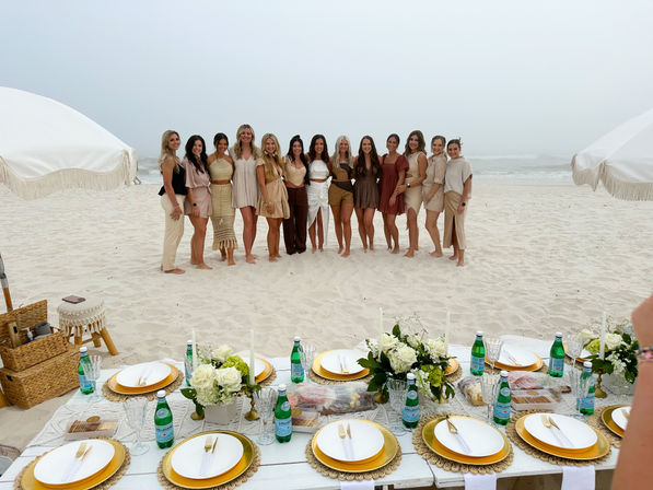 Group of women in neutral outfits posing on a white-sand beach behind a boho-chic picnic setup with gold plates, floral centerpieces, fringe umbrellas — seaside celebration.