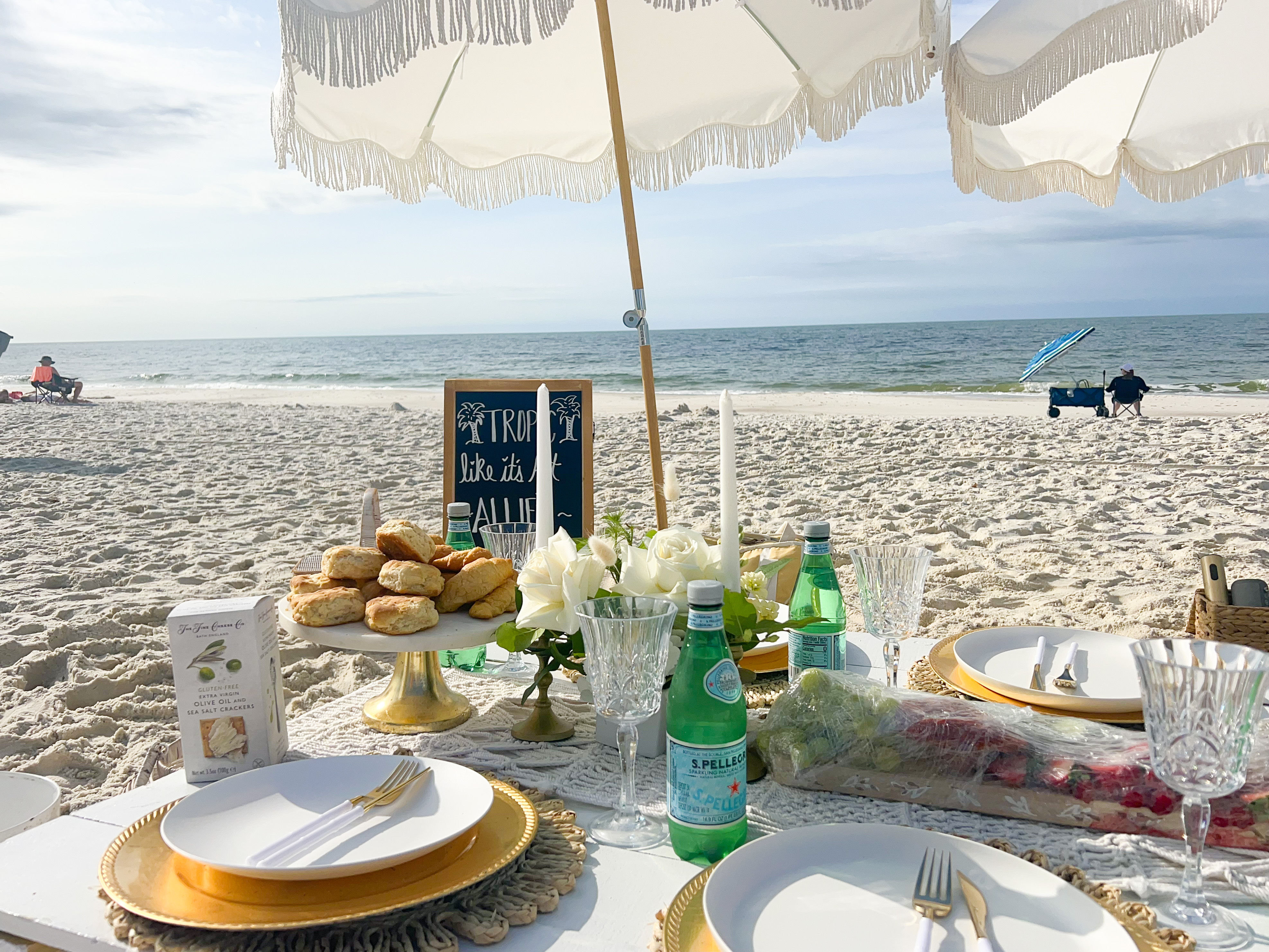 Coastal beach picnic under a white fringed umbrella with gold chargers, crystal glasses, scones on a cake stand, sparkling water bottles and white floral centerpiece on a low table by the sandy shore and ocean.