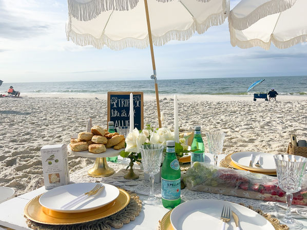 Coastal beach picnic under a white fringed umbrella with gold chargers, crystal glasses, scones on a cake stand, sparkling water bottles and white floral centerpiece on a low table by the sandy shore and ocean.