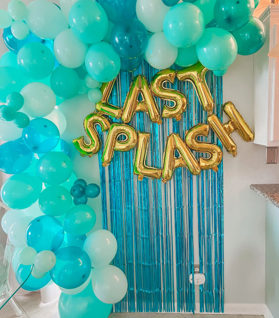 Turquoise and aqua balloon arch framing a blue fringe party backdrop with gold foil letter balloons spelling 'LAST SPLASH' on a home kitchen wall