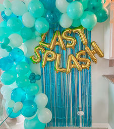 Turquoise and aqua balloon arch framing a blue fringe party backdrop with gold foil letter balloons spelling 'LAST SPLASH' on a home kitchen wall