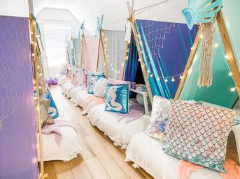 Playful mermaid-themed indoor kids’ sleepover with pastel mini teepee tents, sequin mermaid and scale-print pillows, fairy string lights and decorative netting along a hardwood floor