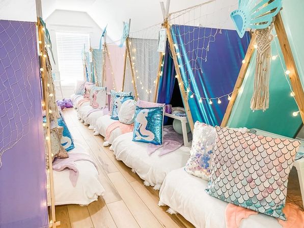 Playful mermaid-themed indoor kids’ sleepover with pastel mini teepee tents, sequin mermaid and scale-print pillows, fairy string lights and decorative netting along a hardwood floor