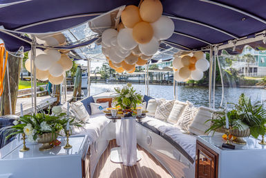 Decorated pontoon boat interior with navy canopy, peach and white balloon clusters, cushioned wraparound seating, floral arrangements and a central table with appetizers, docked along a sunny waterfront canal.