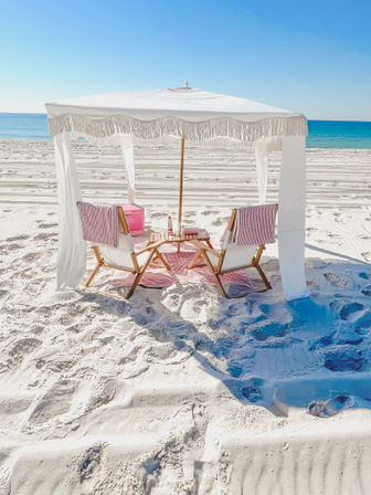 Fringed white beach cabana with two wooden lounge chairs draped in pink-striped towels, a small table and cooler on pristine white sand facing a turquoise ocean under a clear blue sky.