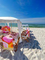White-sand beach with a fringed white cabana, wooden lounge chairs draped in colorful towels, a pink swirl inflatable, and calm turquoise ocean under a bright blue sky — sun-soaked seaside setup.