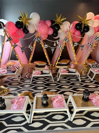 Cozy indoor glam sleepover setup with pink fabric teepees, pink-white-black balloon garlands topped with gold star balloons, fairy lights, low white trays with lanterns and pink napkins, plush pillows on a black-and-white geometric rug