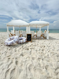 Oceanfront white sandy beach with two fringe-topped white cabanas, wooden sun loungers, patterned inflatable tubes and a small chalkboard sign facing calm turquoise water under a partly cloudy sky.
