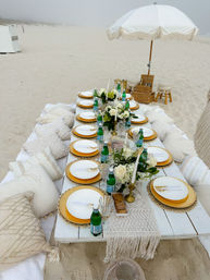 Boho beach picnic on white sand — low wooden table set for a group with gold chargers, white plates, green glass water bottles, floral centerpieces, candles, cushions and a fringed white umbrella on a misty shoreline.