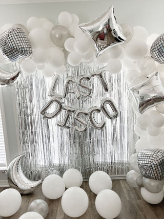Silver-and-white indoor party backdrop with metallic fringe curtain, foil letter balloons spelling LAST DISCO, white and silver balloon arch, mirrored disco ball and star accents, scattered floor balloons