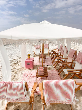 White fringed beach cabana sheltering wooden lounge chairs draped in pink-and-white striped towels, pink cooler and picnic boxes on white-sand shore under a pale blue sky.