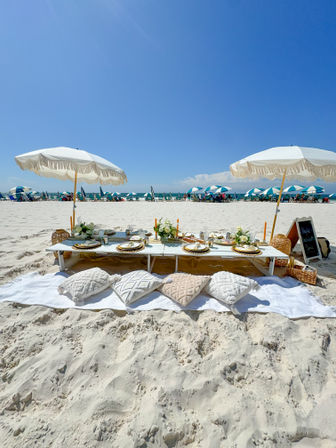Sunny white-sand beach picnic with low wooden table, textured pillows, fringe umbrellas, place settings and flowers facing turquoise ocean under a clear blue sky