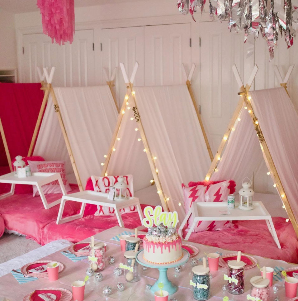 Cozy pink indoor kids' slumber party setup with row of pale-pink teepee tents wrapped in fairy lights, plush cushions and low white trays; foreground table with pink drip cake on blue stand, paper cups, jars of sweets and disco-ball decorations.