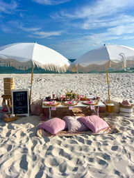 Sunny beach picnic setup on white sand with two fringed white umbrellas, low table with floral centerpieces and place settings, pink floor cushions, a chalkboard sign reading "Happy Sweet 16," and blue ocean background.
