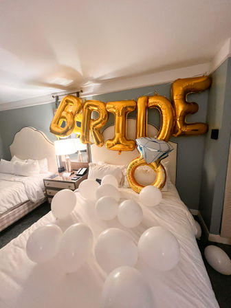 Hotel room decorated for a bridal celebration with gold foil "BRIDE" and oversized diamond ring balloons above a bed strewn with white balloons