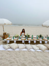 Woman sitting at a boho beach picnic on white sand with a low table set for dining—gold-rimmed plates, green glass bottles, floral centerpieces, fringe umbrellas and cozy pillows, foggy ocean waves in the background