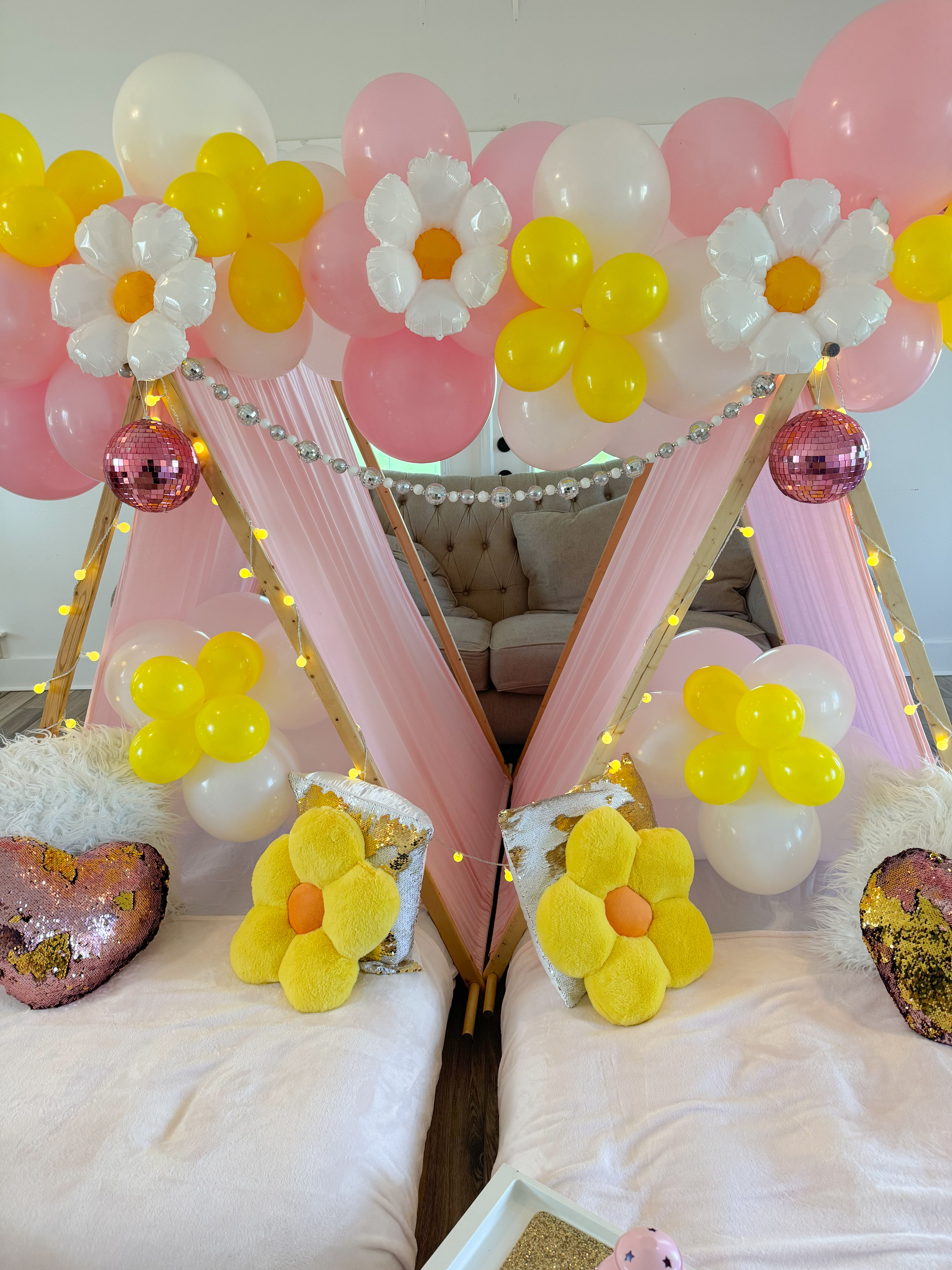 Indoor kids' slumber party setup in a living room with two pink teepee tents, pink/white/yellow balloon garlands and daisy balloon accents, string lights, pink disco balls, and plush yellow flower pillows on cozy mattresses.
