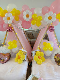 Indoor kids' slumber party setup in a living room with two pink teepee tents, pink/white/yellow balloon garlands and daisy balloon accents, string lights, pink disco balls, and plush yellow flower pillows on cozy mattresses.
