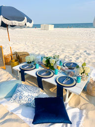 Sunlit white-sand beach picnic with a low white table, navy-blue plates and napkins, floral centerpiece, cushions and fringed umbrella by the ocean