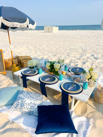 Sunlit white-sand beach picnic with a low white table, navy-blue plates and napkins, floral centerpiece, cushions and fringed umbrella by the ocean