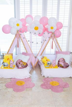 Indoor kids' slumber party setup with two pink mini teepees by shuttered windows, pink and white balloon garland with daisy-shaped balloons, plush pillows (yellow flower, fluffy white, sequin heart), trays with disco balls and lanterns, and pink flower-shaped rugs on tile floor.