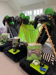 Gaming-themed kids' birthday sleepover in a home: green-and-black mini teepees with string lights, inflatable controller balloons, gamepad pillows, floor cushions and trays with controllers and headsets.