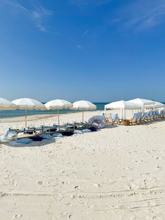 White-fringed beach umbrellas and low tables with cushions set up on powdery white sand beside a calm turquoise ocean under a clear blue sky.