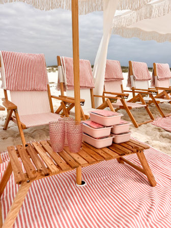 White-sand beach setup with wooden lounge chairs draped in pink-and-white striped towels under a fringe umbrella, small wooden table holding stacked pink meal containers and textured pink cups, cloudy coastal sky