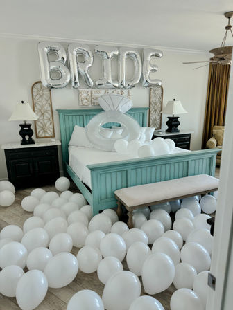 Bridal bedroom decorated for a bachelorette celebration: turquoise bed with giant silver "BRIDE" balloons overhead, oversized inflatable ring on the bed and dozens of white balloons scattered across the floor.