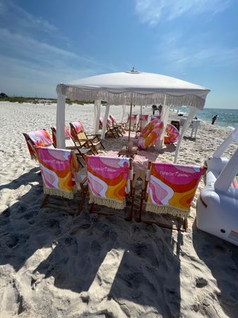 Sunlit beach scene with a white fringed canopy shading wooden lounge chairs draped in colorful retro towels, inflatable floaties on white sand and the turquoise ocean under a clear blue sky.