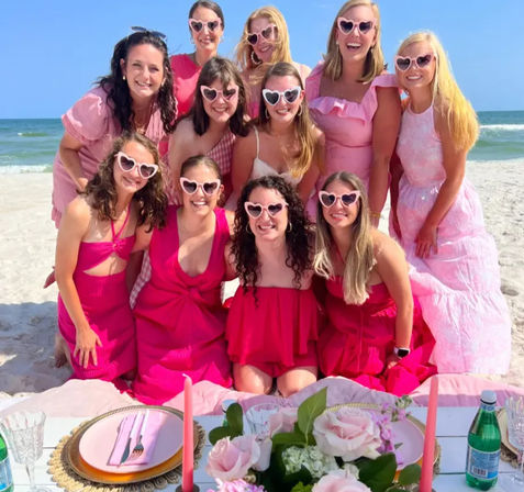 Group of friends in pink dresses and heart-shaped sunglasses posing on a sunny beach behind a pink picnic setup with roses, candles and ocean waves in the background.
