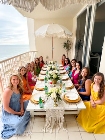 Group of friends in colorful sundresses enjoying a seaside balcony dinner party with a long white table, gold chargers, floral centerpieces and an ocean view under a fringed umbrella.