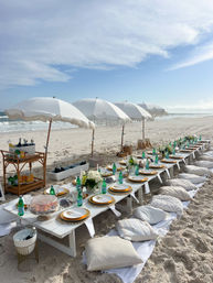 Coastal-chic beach dining on white sand: long low tables with fringe umbrellas, cushions for floor seating, floral centerpieces and green glass water bottles by the ocean under a blue sky.