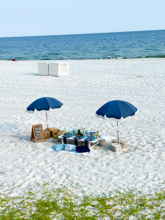 Seaside picnic on white sand: low table set with blue plates, cushions and floral decor under two navy umbrellas with calm blue ocean beyond.