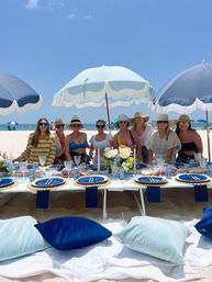 Group of friends at a styled beach picnic on white sand, blue fringe umbrellas overhead, low table set with navy plates, candles and floral centerpieces, cushions and the ocean in the background