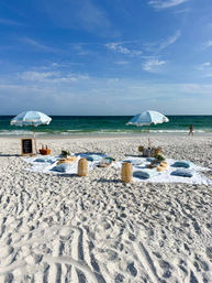 Sunlit white-sand beach picnic with two pale-blue umbrellas, floor cushions, wicker lanterns and turquoise ocean under a bright blue sky.