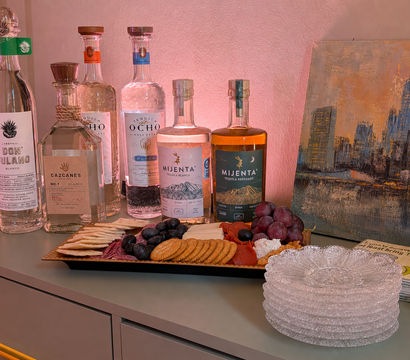 Inviting tequila tasting display on a gray sideboard: clear and amber tequila bottles, charcuterie platter with crackers, grapes, olives, cheeses and cured meats, stacked glass plates and cityscape artwork under warm pink lighting