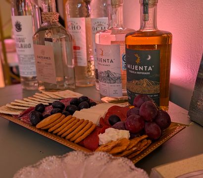 Charcuterie platter with crackers, sliced salami, cheeses, black olives and red grapes on a tray, set in front of several tequila bottles on a pink-lit indoor bar countertop, ready for tasting.
