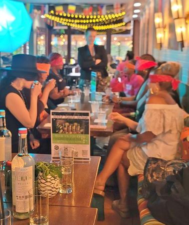 Blindfolded cocktail tasting at a lively restaurant bar — guests in colorful blindfolds seated along a long table, mezcal bottle and string lights overhead.