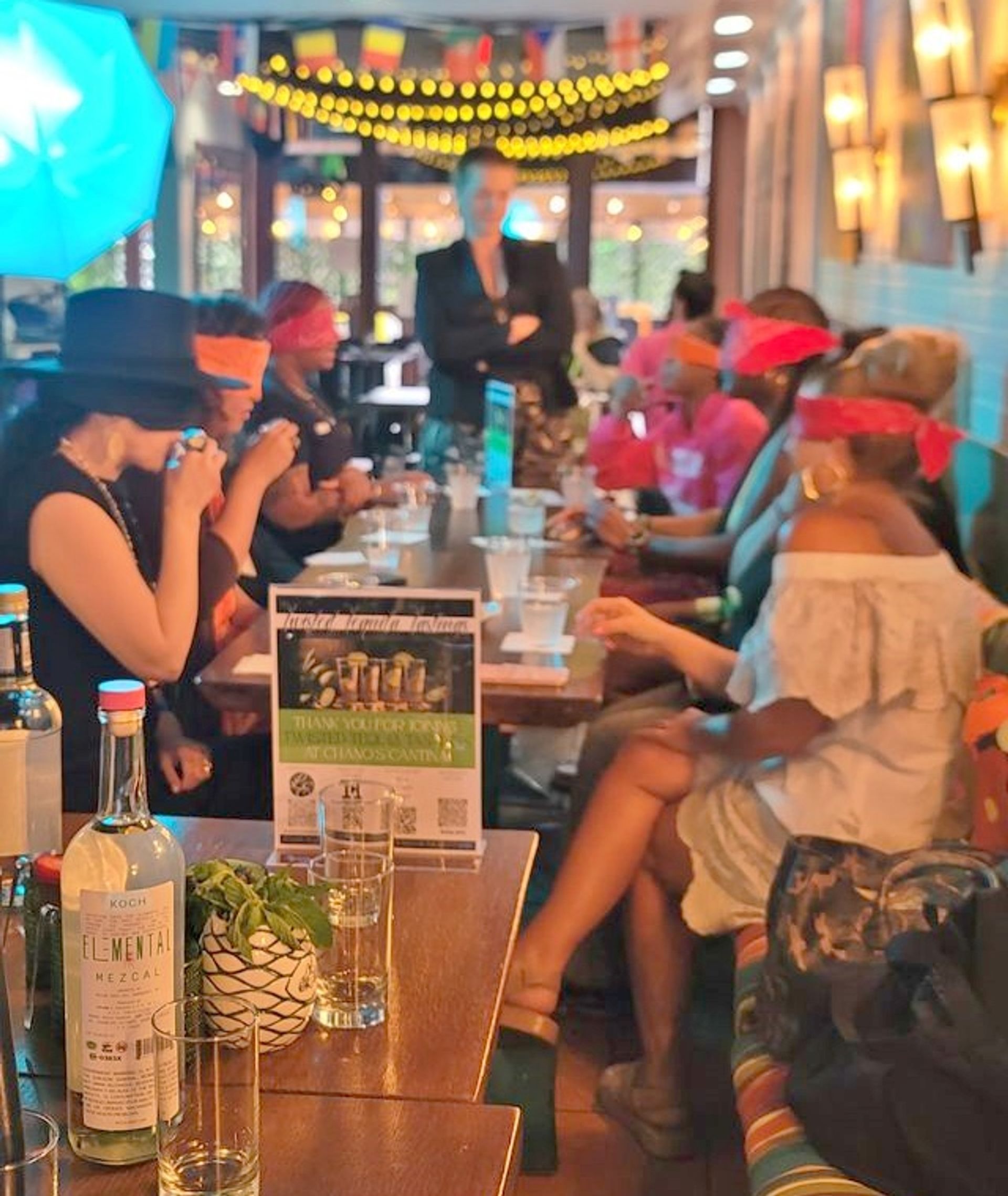 Blindfolded cocktail tasting at a lively restaurant bar — guests in colorful blindfolds seated along a long table, mezcal bottle and string lights overhead.
