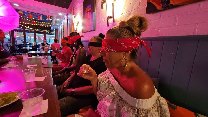 Blindfolded group in red bandanas seated along a neon-lit cocktail bar taking shots; long wooden table with shot glasses, lime slices, plastic cups, string lights and colorful wall art