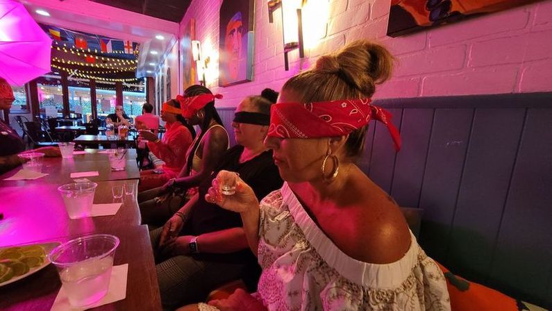 Blindfolded group in red bandanas seated along a neon-lit cocktail bar taking shots; long wooden table with shot glasses, lime slices, plastic cups, string lights and colorful wall art