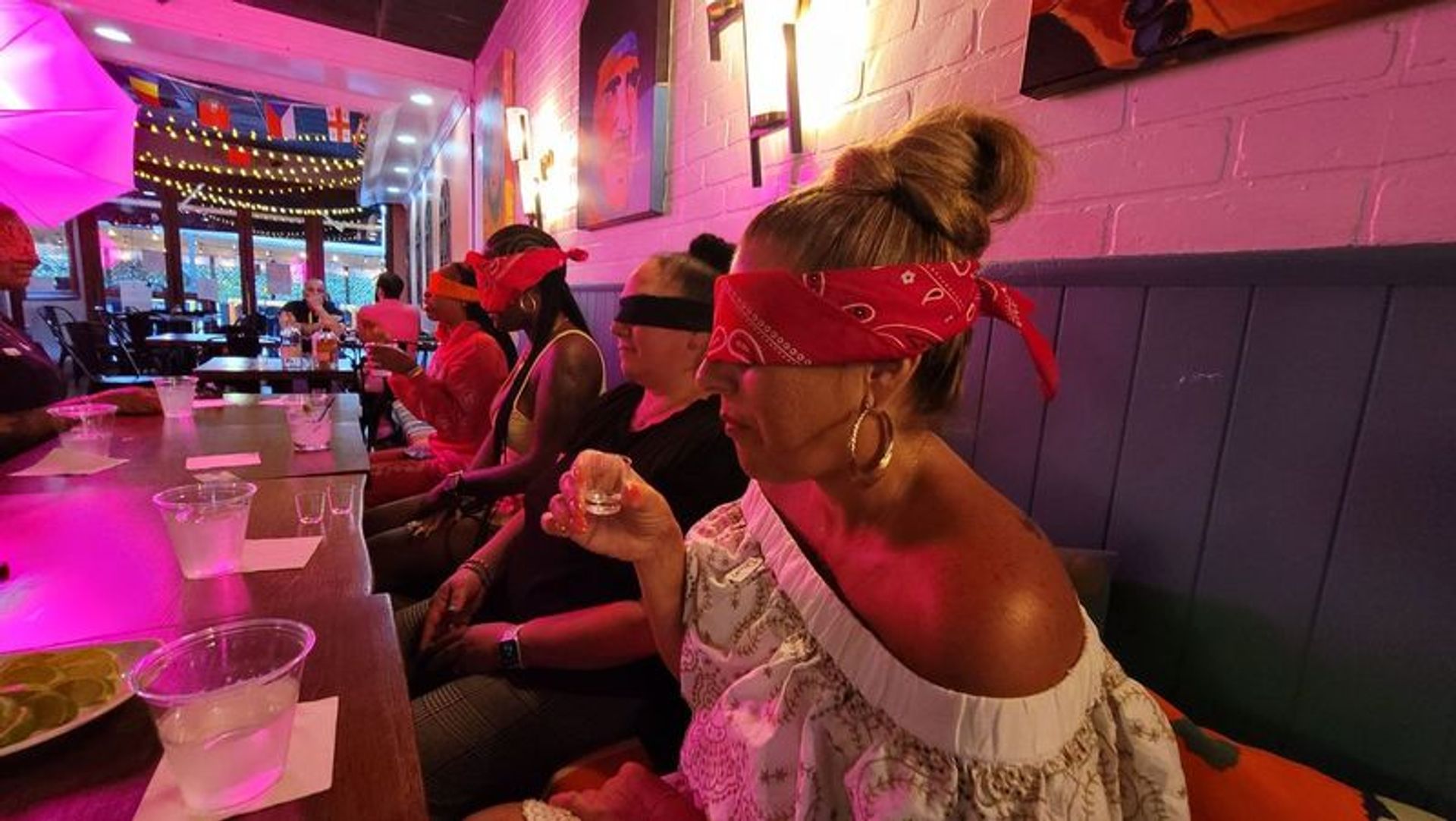 Blindfolded group in red bandanas seated along a neon-lit cocktail bar taking shots; long wooden table with shot glasses, lime slices, plastic cups, string lights and colorful wall art