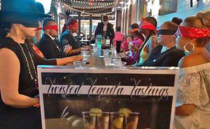 Blindfolded adults lined up along a long bar under string lights, sampling shots and guided by a server during a lively indoor tequila tasting event.